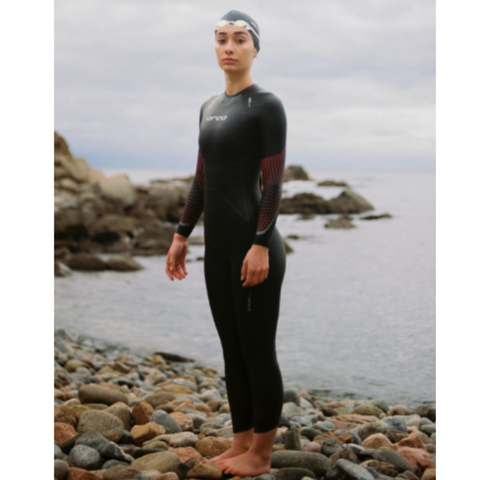 A woman wearing a full-length black wetsuit with red patterned arm panels and a white swim cap stands on a rocky shoreline with water and rocks in the background.