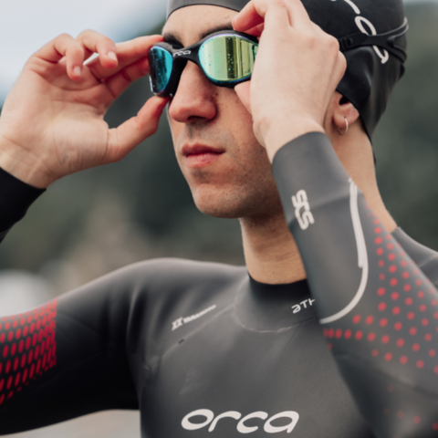 A person wearing openwater swimming hat, goggles and wetsuit. A white Orca logo is visible on each item. Set against a blurred out background.