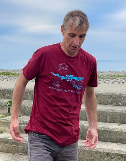 A person wearing a burgundy t-shirt with a baby blue logo and graphic walking outdoors with a blurred background.