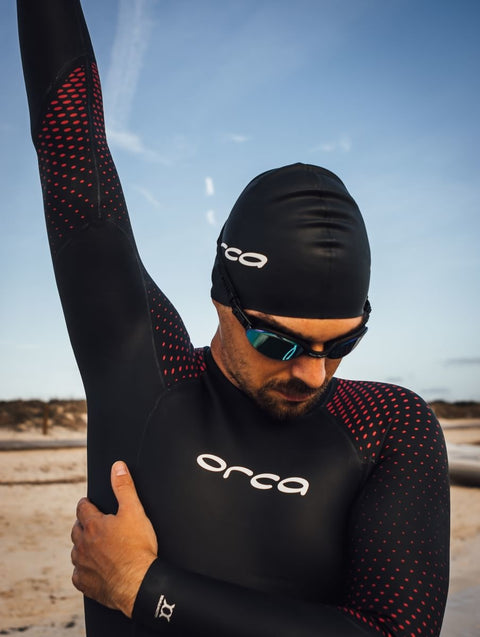 A picture a of a man wearing a black and red wetsuit, goggles and a black swimming hat stood on a beach.